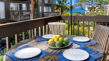 A wooden table set with plates, a bowl of fruit, and a blue woven placemat on a balcony overlooking a residential area with a view of the ocean.