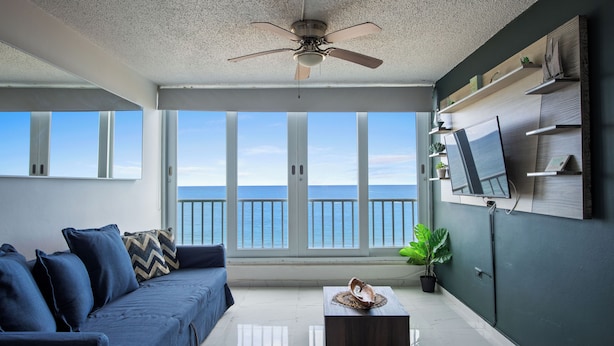 A modern living room with a large window overlooking the sea, a ceiling fan, a sofa, a coffee table, and a mounted TV. - Marina Beach 2 Bd Oceanfront Pool Parking (Carolina)