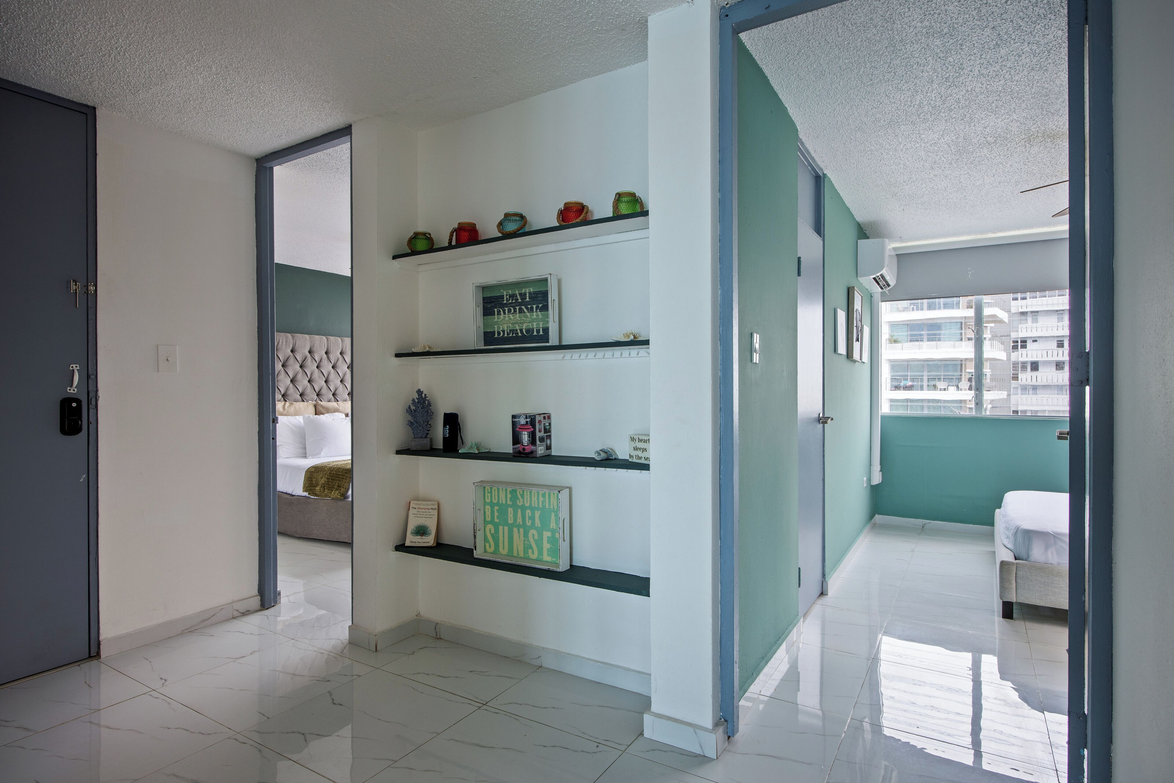 A hotel room with a bed, a shelf with decorative items, a door, and a view of the outside.