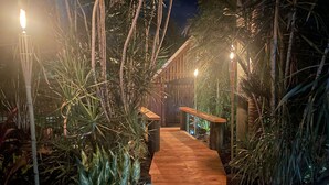 Un sendero de madera bordeado por exuberante vegetación tropical, que conduce a una estructura de madera con un banco.