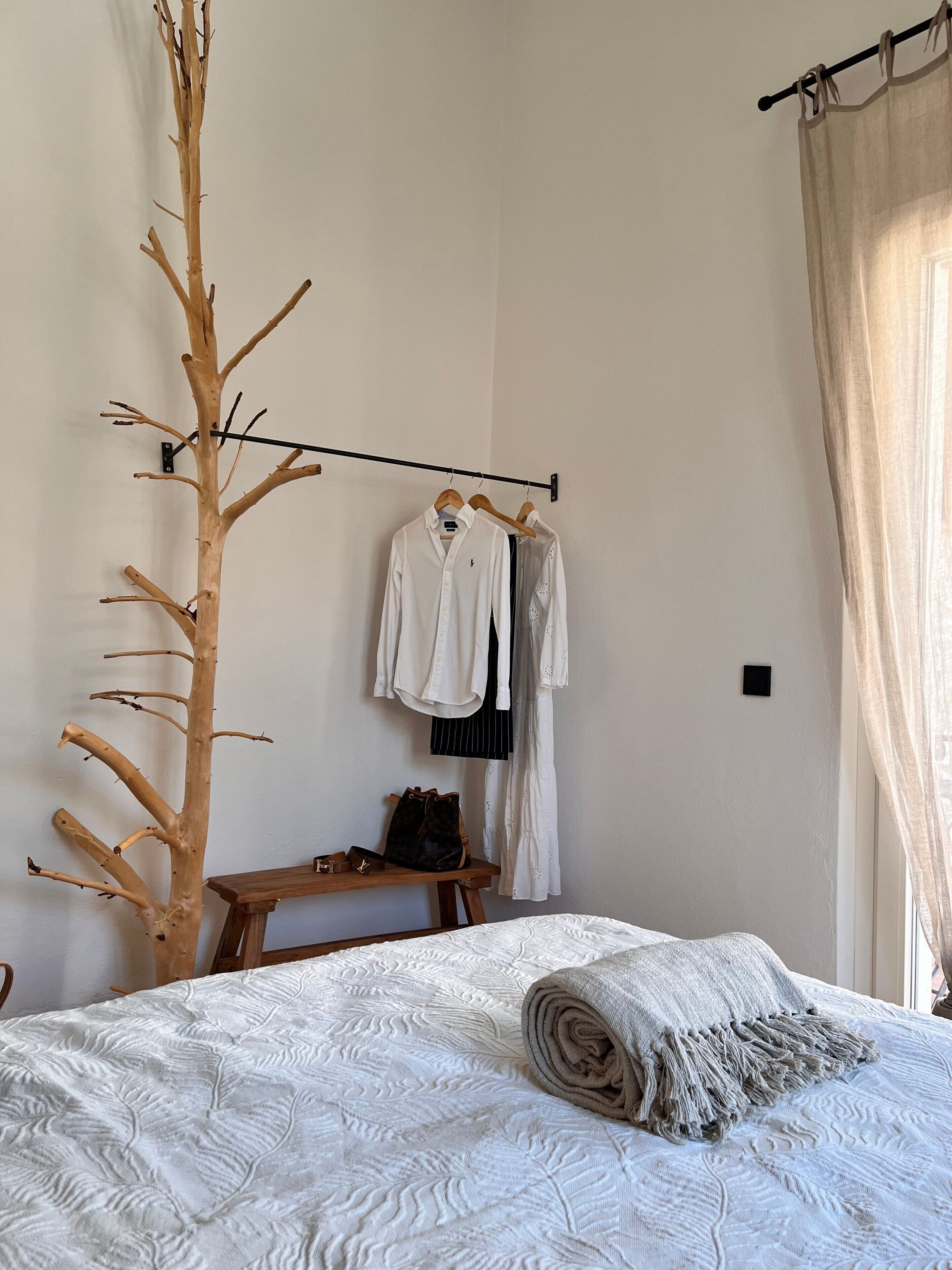 Ein Schlafzimmer mit einem Bett, einer Wanddekoration aus Holz, einem Kleiderständer mit Kleiderbügeln, einer Bank und einem Fenster mit Vorhängen.