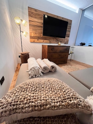 Une chambre moderne avec un grand lit, une commode en bois, un téléviseur à écran plat fixé sur un panneau en bois et une lampe de chevet.