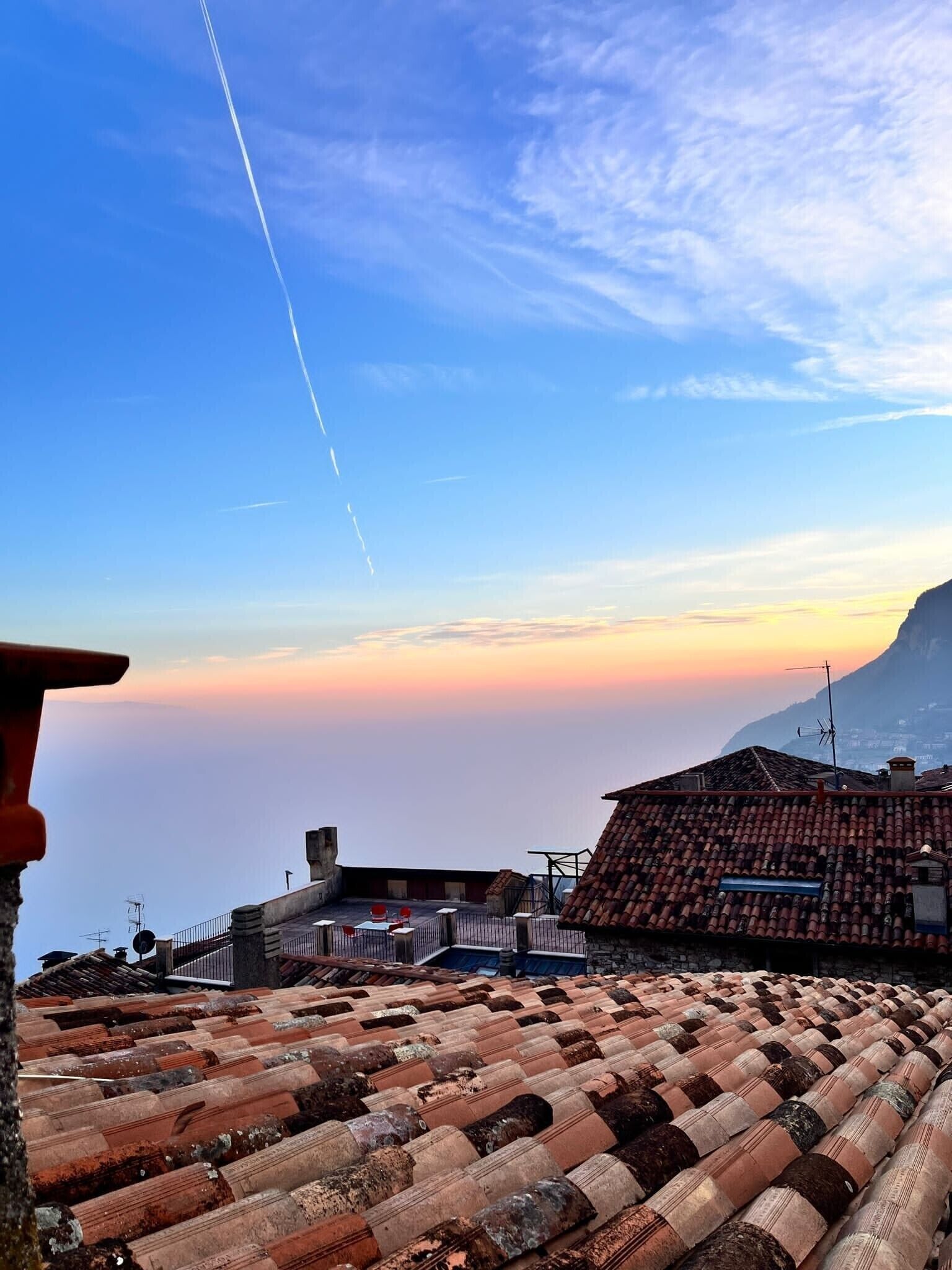 Un toit recouvert de tuiles en terre cuite, un ciel dégagé et une montagne lointaine.