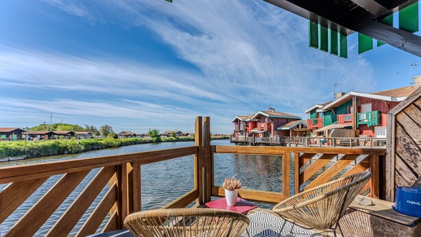 A wooden deck with wicker chairs and a table, overlooking a river with houses in the background.