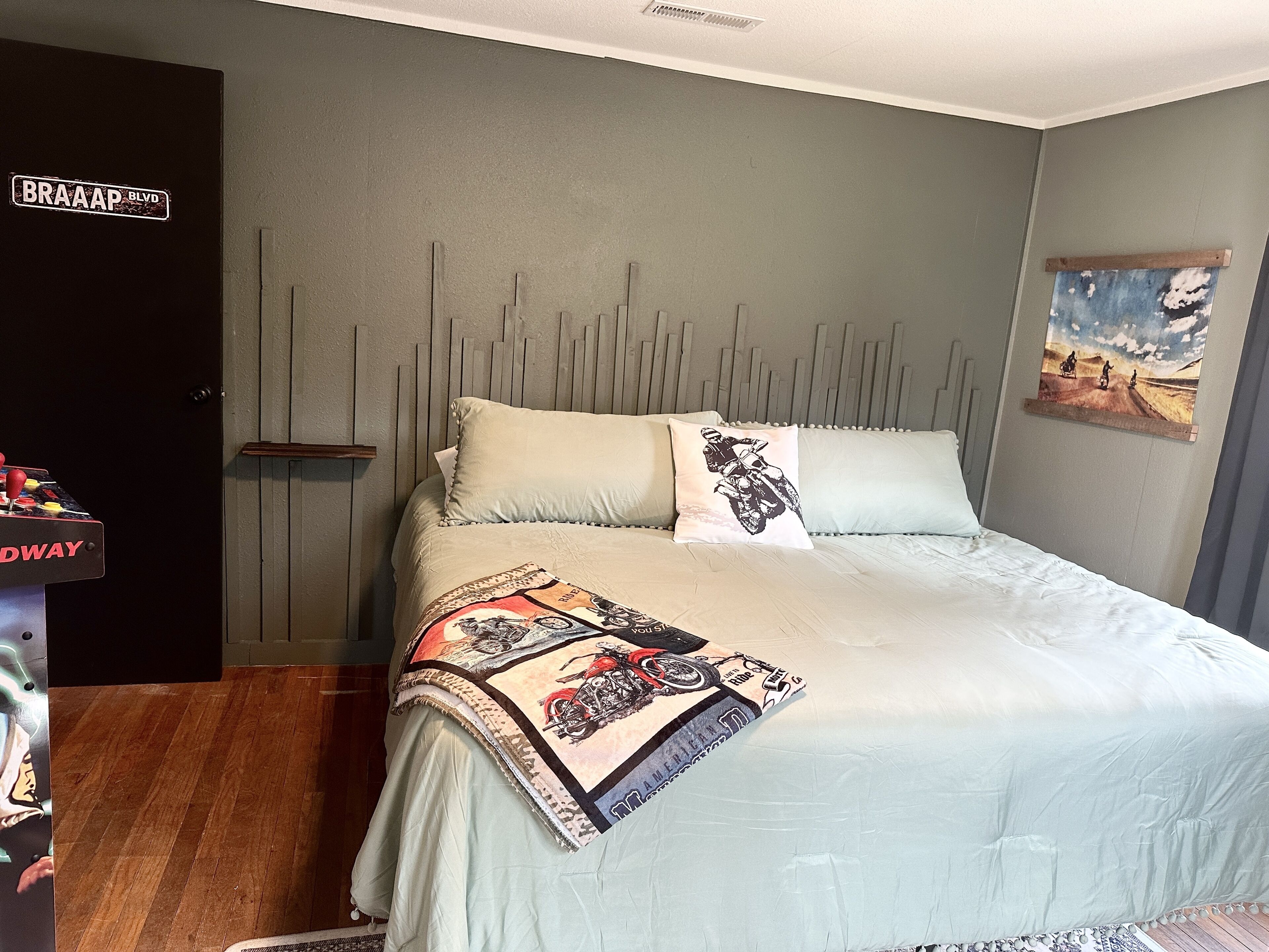 A bedroom with a bed, pillows, a blanket, a wall-mounted artwork, and a door with a sign.