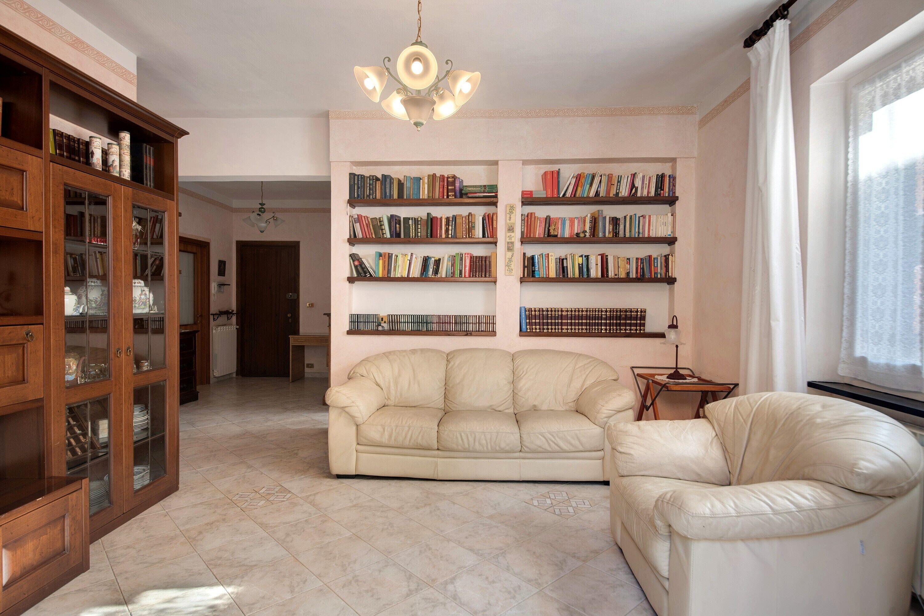 A living room with a beige leather sofa, a wooden bookcase with books, a chandelier, and a window with curtains.