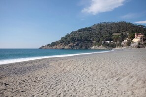 Una spiaggia con ciottoli, una collina con alberi e edifici, e un cielo azzurro limpido.