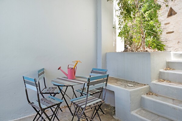 A small outdoor seating area with a table and chairs, a pink watering can, and a staircase leading to a higher level.