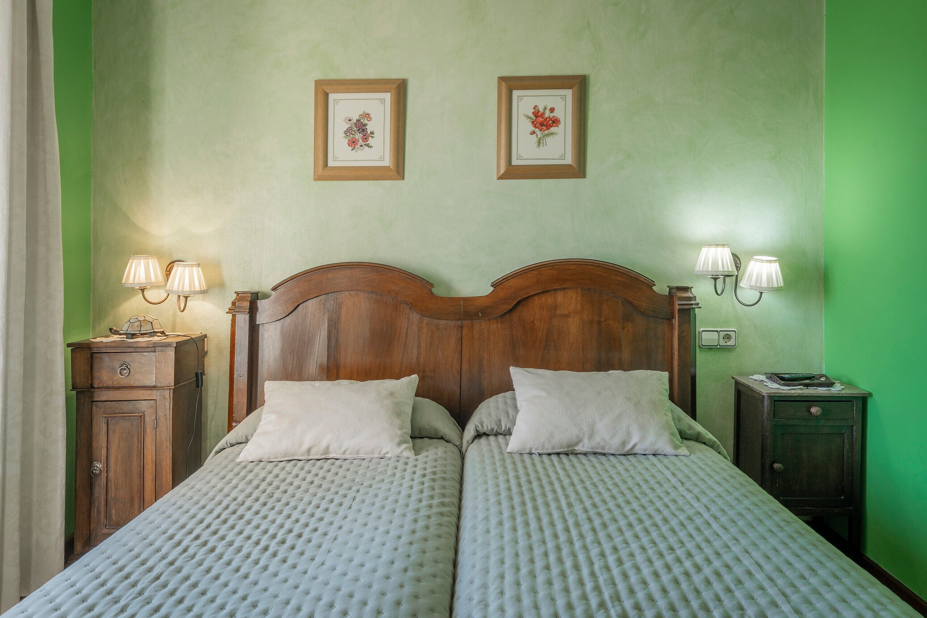 Een tweepersoonsbed met een houten hoofdeinde, twee nachtkastjes met lampen en twee ingelijste bloemenprints aan de muur.