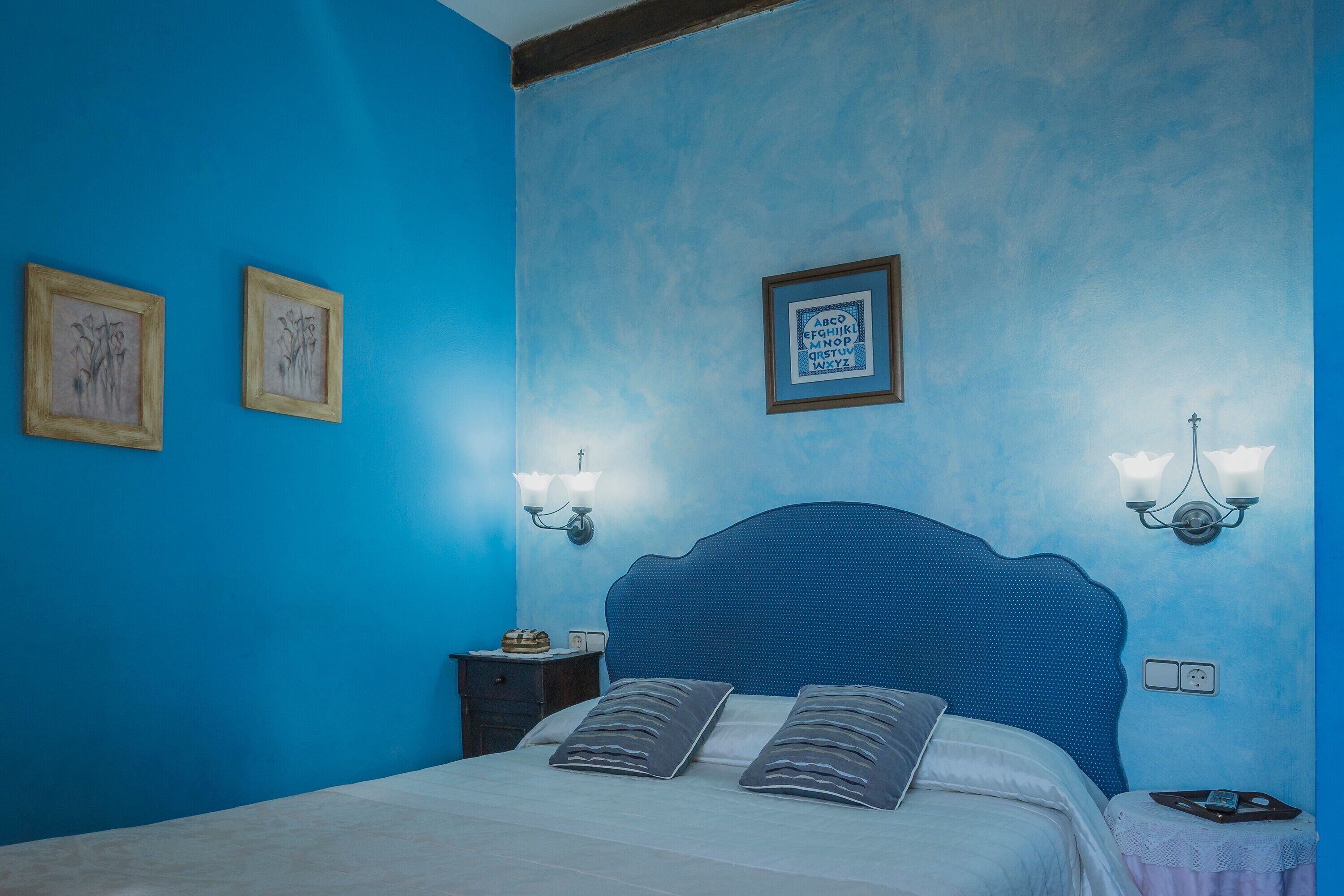 A bedroom with a blue wall, a bed with two pillows, a nightstand with a lamp, and framed pictures on the wall.