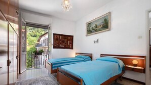 Ein Schlafzimmer mit zwei Betten, einem hölzernen Kopfteil, einem Nachttisch, einer Chandelieber, einem Gemälde und Blick auf einen Balkon mit einem Haus.