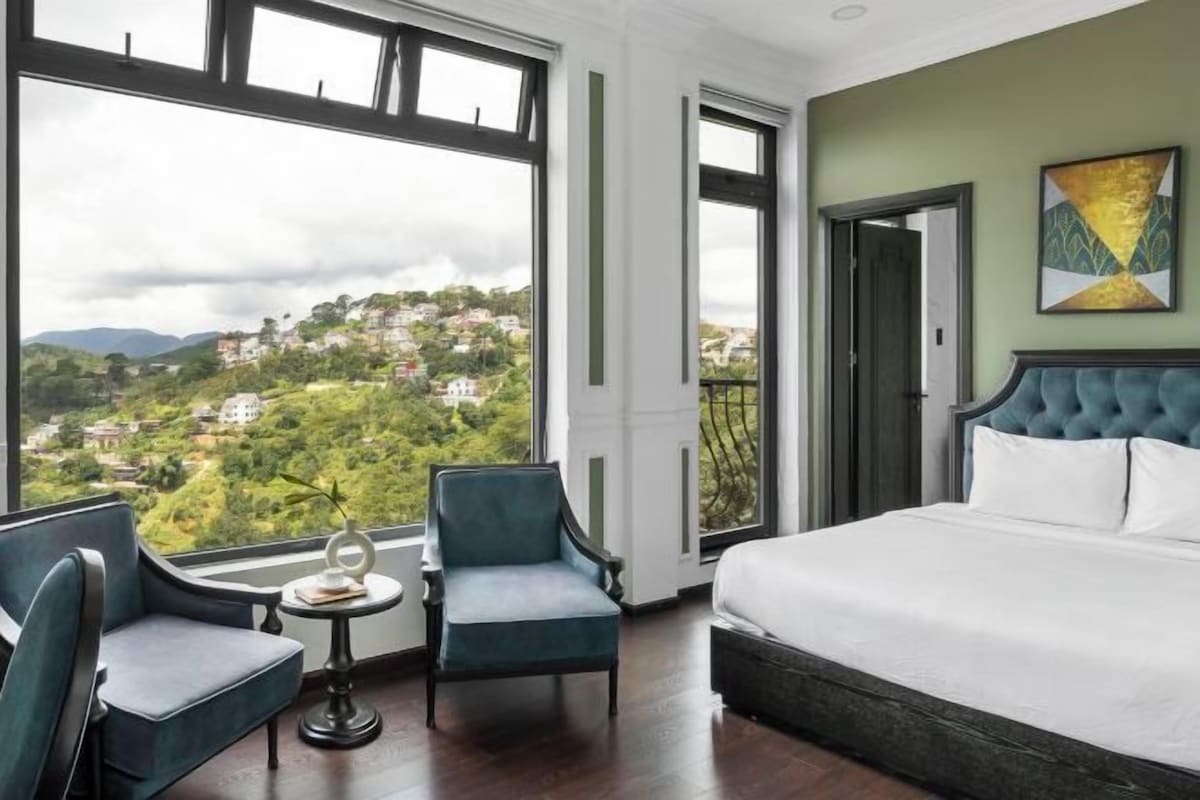 Habitación de hotel con una cama grande, dos sillones, una mesita y vistas a un pueblo en la ladera a través de amplias ventanas.