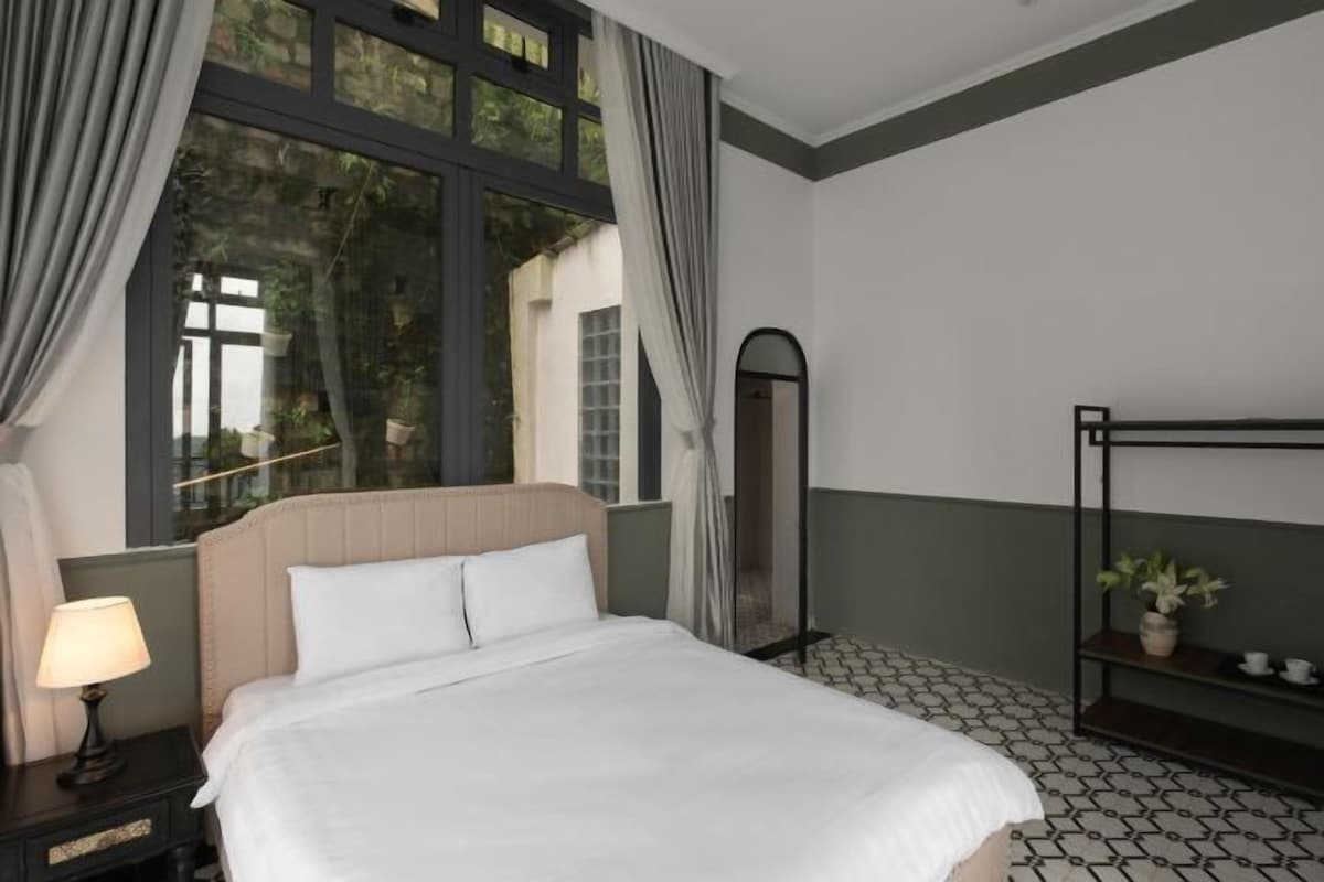 Habitación de hotel con una cama, una mesita de noche con lámpara, un espejo, una estantería con plantas y una ventana con cortinas.
