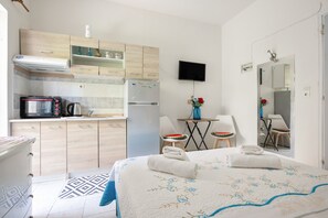 Habitación de hotel compacta con kitchenette, una cama con cubrecama floral, una pequeña mesa de comedor y un televisor montado en la pared.