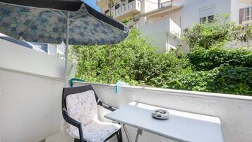 A balcony with a white table and chair, a patterned umbrella, and a view of a white building and greenery.