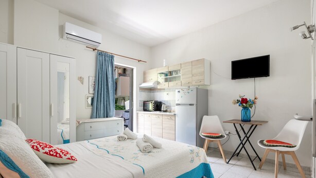 A bedroom with a bed, a desk, a chair, a TV, and a kitchenette.
