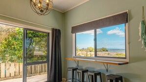 A room with a large sliding glass door leading to an outdoor deck, a window with a view of the landscape, and three bar stools. - Sleeps 4, Estuary Front in Pukehina Beach, two minutes to beach (Pukehina)