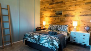 A bedroom with a wooden wall, a bed with a tropical print bedspread, two bedside lamps, a nightstand, and a globe on the nightstand. - Sleeps 4, Estuary Front in Pukehina Beach, two minutes to beach (Pukehina)