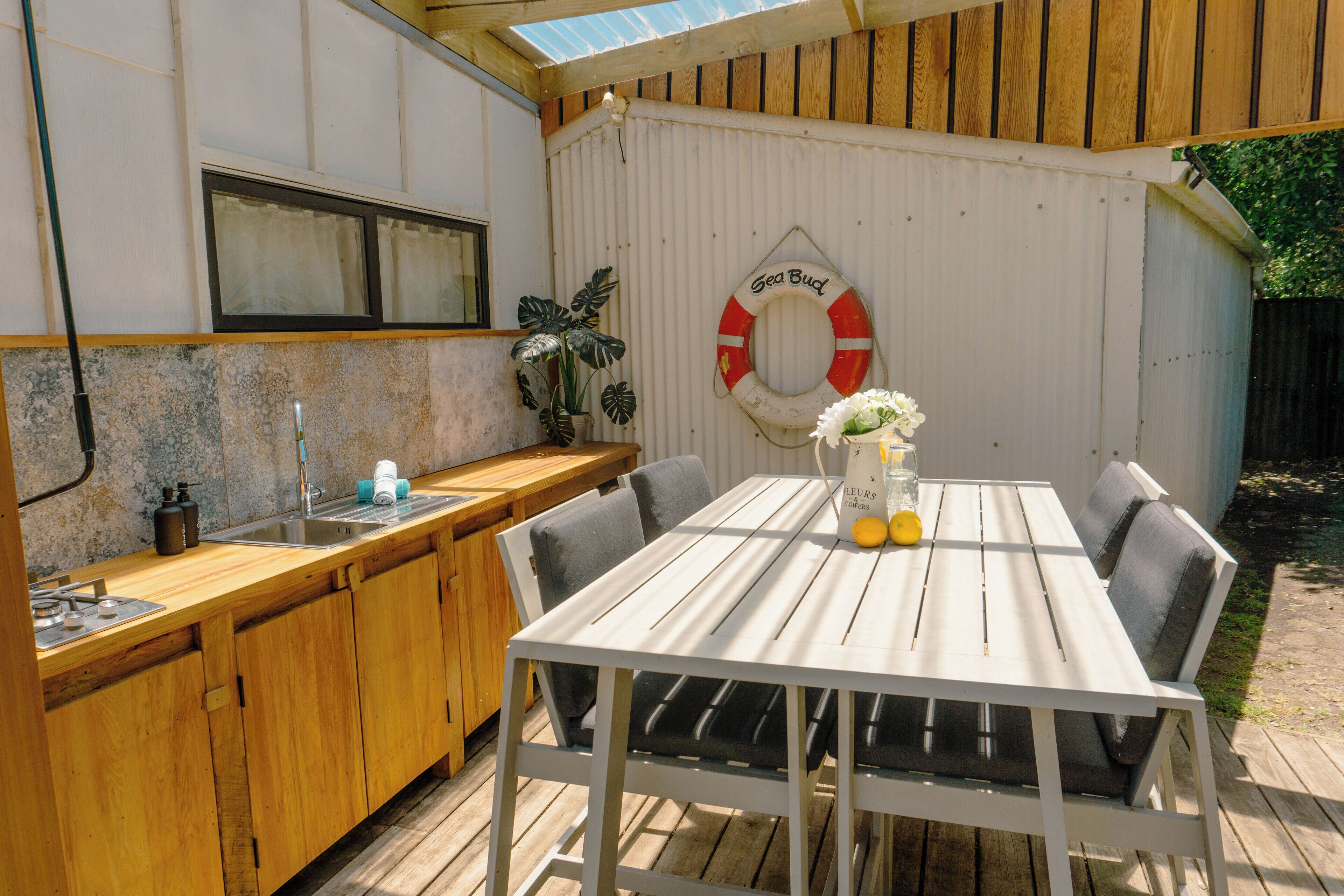 Eine Holzterrasse mit einem weißen Tisch und Stühlen, ein kleiner Küchenbereich mit Spüle und Schränken sowie ein Rettungsring an der Wand.