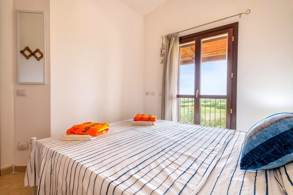 A bedroom with a bed, a window with curtains, a mirror, and a view of greenery outside. - Holiday Home 'La Capazza' with Sea View, Wi-Fi and Air Conditioning (Viddalba)