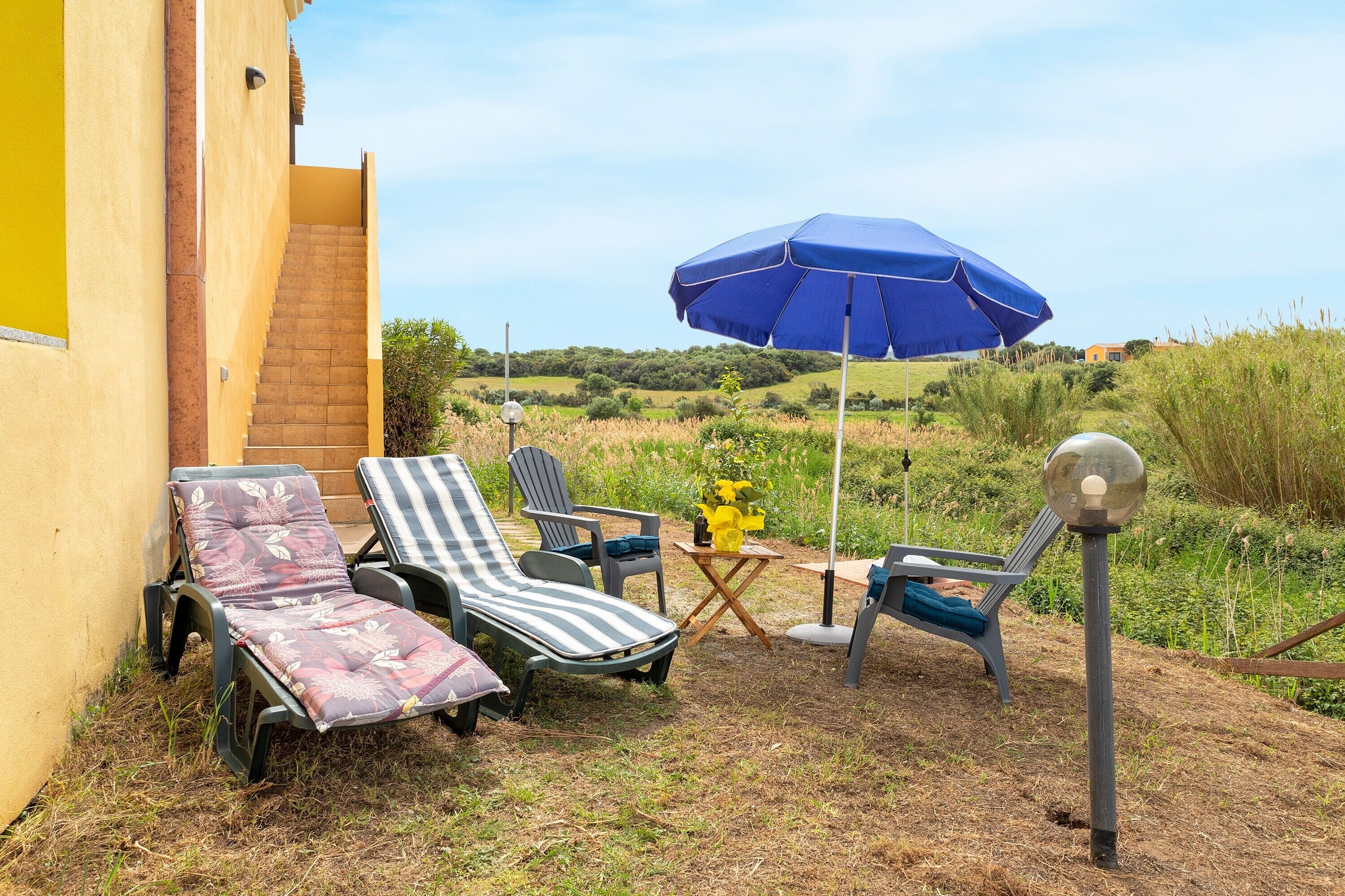 A patio with two lounge chairs, a blue umbrella, and a small table with a vase of flowers.