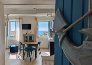 A dining area with a wooden table and chairs, a television mounted on the wall, and a view of the sea through large windows.