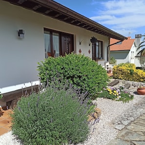 Ett hus med vit fasad, en träveranda och en trädgård med lavendel och gula blommor.