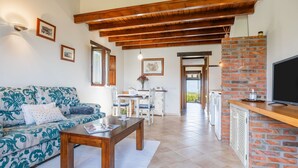 Uma sala de estar com sofá, mesa de centro e lareira. Há uma área de jantar com cadeiras e mesa. O quarto tem vigas de madeira no teto e uma parede de tijolos.