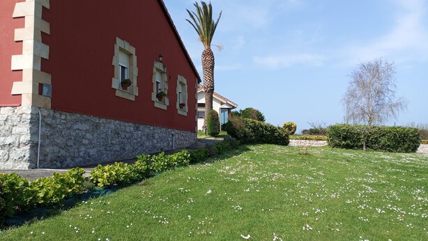 Un edificio rojo con base de piedra, una palmera y un césped bien cuidado con flores blancas.