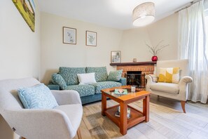 Ein Wohnzimmer mit Sofa, Sessel, Couchtisch, Kamin und gerahmten Pflanzenbildern an der Wand.