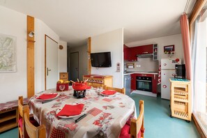 Cozinha compacta com armários vermelhos, uma TV fixada na parede e um espaço de refeições com uma mesa posta para dois.
