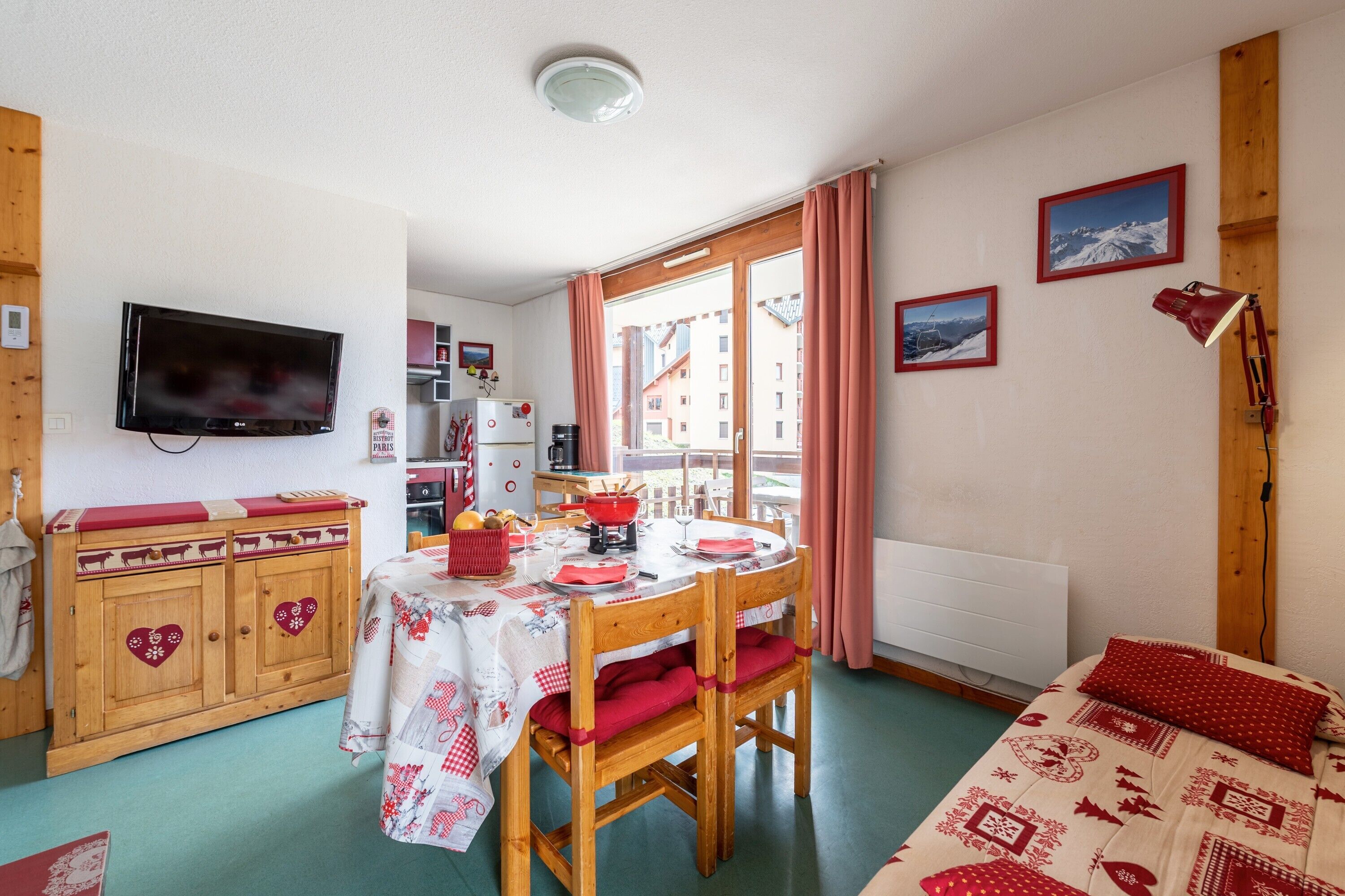 Une pièce chaleureuse avec une table à manger en bois, des chaises, un lit avec une literie rouge, un téléviseur fixé au mur et une fenêtre avec des rideaux.