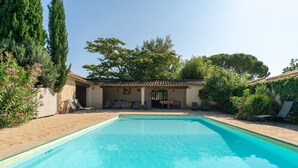 A swimming pool with a tiled deck, a covered seating area, and surrounding greenery.