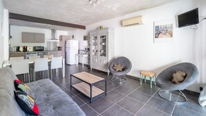 A modern living room with a grey sofa, a wooden coffee table, a grey armchair, and a dining area with white chairs and a kitchen in the background.