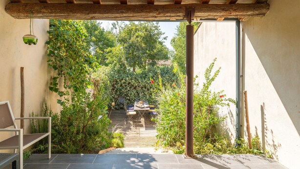 A covered patio with a table and chairs, surrounded by greenery and a view of a building in the distance.