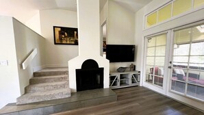 A spacious living area with a staircase, a flat-screen TV, and large windows with blinds.