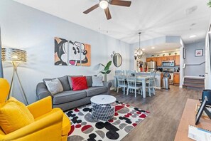 A modern living room with a sofa, a yellow armchair, a dining area with a table and chairs, and a kitchen in the background.
