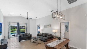 A modern living room with a sofa, coffee table, and dining table. There is a large window with curtains, a ceiling fan, and a hanging light fixture. - Stunning Newly Renovated Pool Home close to Disney (Davenport)