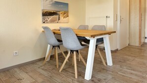 Uma sala de jantar com uma mesa de madeira, quatro cadeiras cinzas, um lustre e um quadro com tema praiano na parede.