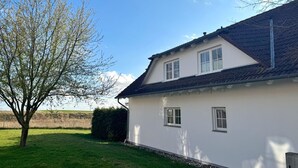 Uma casa branca com telhado inclinado, duas janelas e uma árvore em primeiro plano.