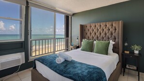 Un dormitorio con un amplio ventanal con vistas a una playa, un cabecero capitonado y una cama bien hecha con una colcha azul y almohadas blancas.