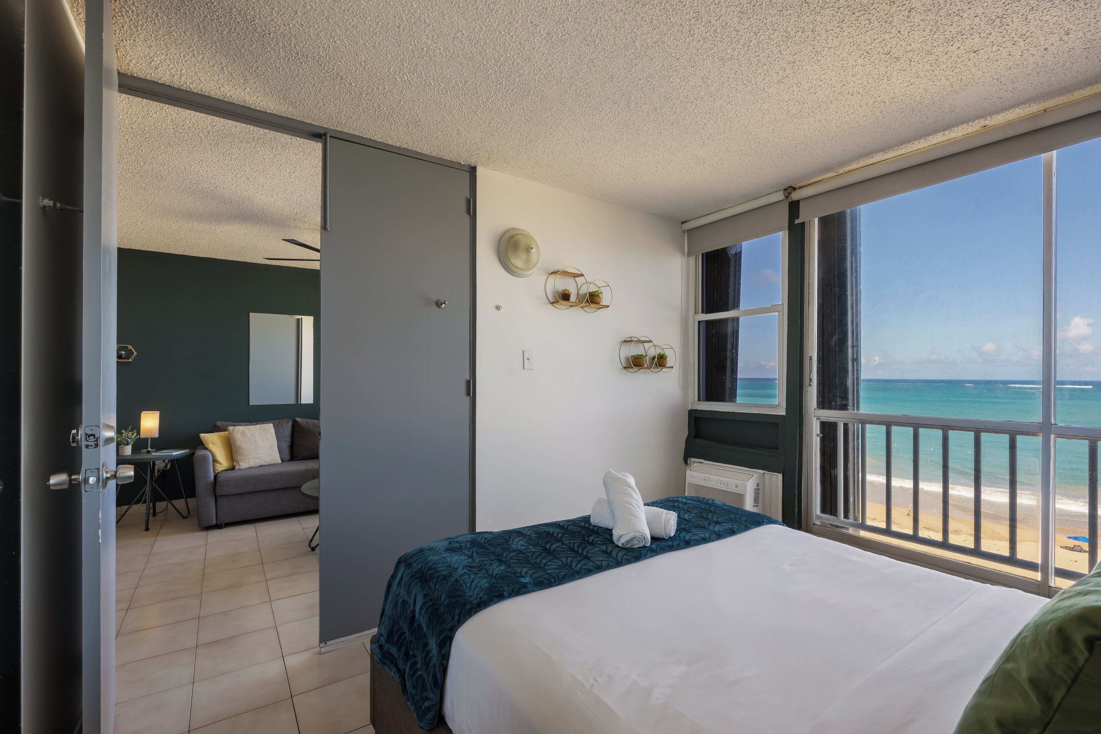 A hotel room with a bed, a view of the beach, a small table with a lamp, and a sofa in the adjacent room.