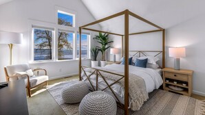A bedroom with a four-poster bed, a view of the sea, a chair, a bedside table with a lamp, and a nightstand with a lamp. - The Perch | Birch Bay Views · Fire Deck · Beach (Blaine)