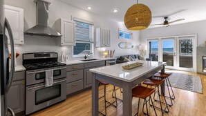 A modern kitchen with stainless steel appliances, a central island with stools, and a view to the outside. - The Perch | Birch Bay Views · Fire Deck · Beach (Blaine)