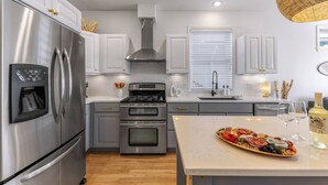 A modern kitchen with stainless steel appliances, a built-in oven, and a central island with a charcuterie board. - The Perch | Birch Bay Views · Fire Deck · Beach (Blaine)