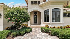 A Mediterranean-style house with a central entrance, a small porch, and a garden with various plants. - 3BR Ritzy Marco Home l Lush Heated Pool (Marco Island)