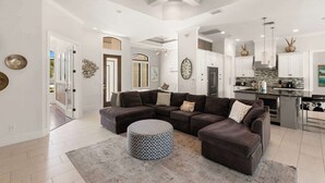 A spacious living room with a sectional sofa, ottoman, and a kitchen with modern appliances. - 3BR Ritzy Marco Home l Lush Heated Pool (Marco Island)