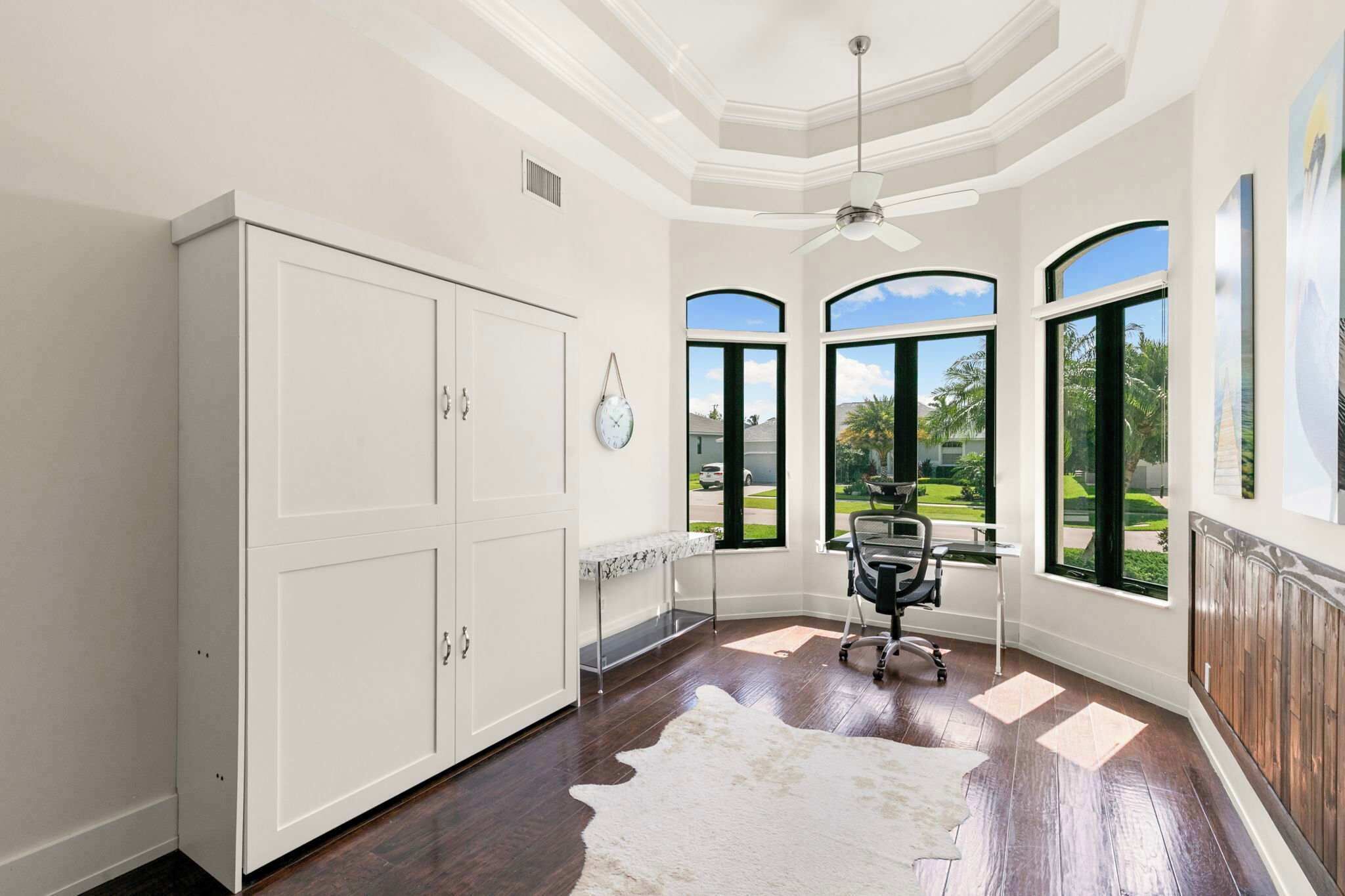 A room with a large white wardrobe, a desk with a chair, a ceiling fan, and large windows with a view of greenery.