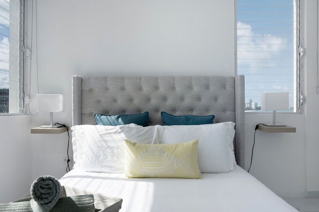 A neatly made bed with white and teal pillows, a grey tufted headboard, and a view of the city through large windows.