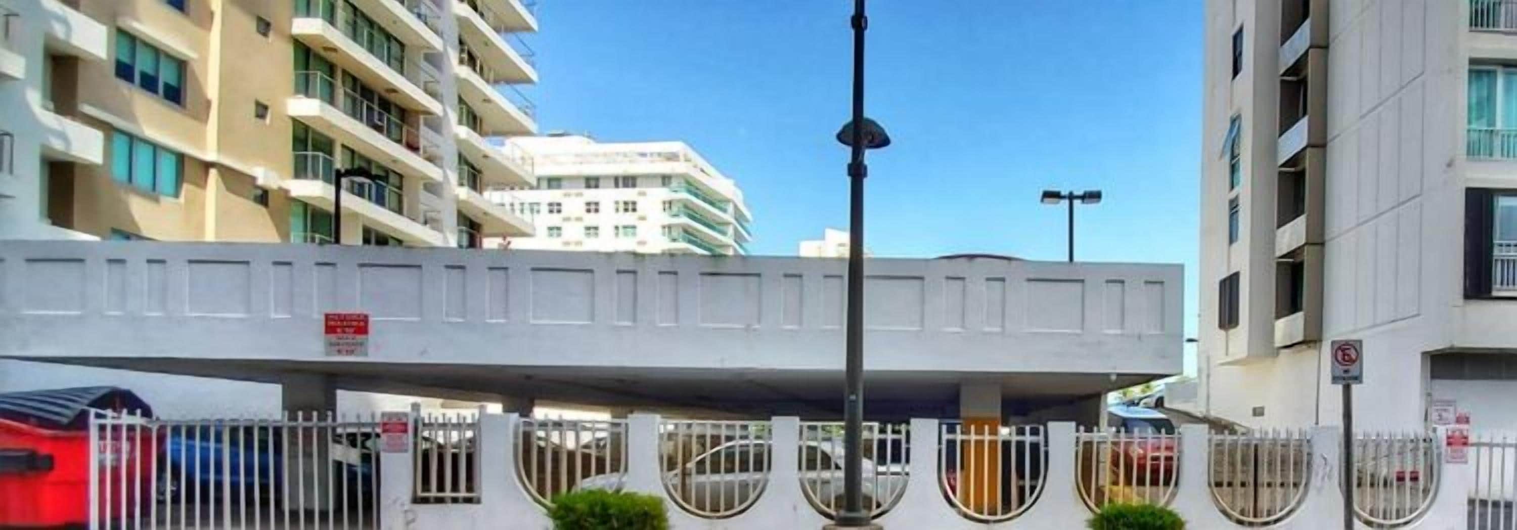 A multi-story building with balconies, a pedestrian bridge, and a gated parking area.